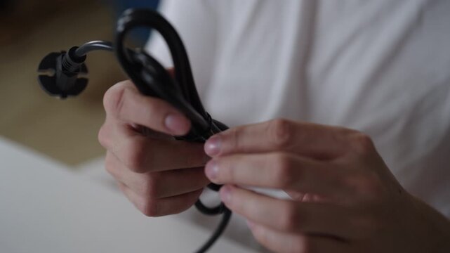 Person hands untangling coiled black electrical power cord, using small black twist tie to secure and organize cable, demonstrating wire management and organization, close-up. Shooting in slow motion.