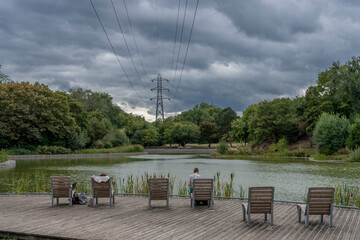 Gennevilliers, France - 08 30 2025: Chanteraines park. People sitting on wooden armchairs in front of the artificial pond  and an electricity pylon in the background with trees and cloudscape