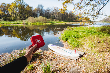 Prioritizing Safety First A hand holding a comprehensive First Aid Kit positioned near a calm and serene river while enjoying paddle boarding. Perfect for an adventurous autumn outing