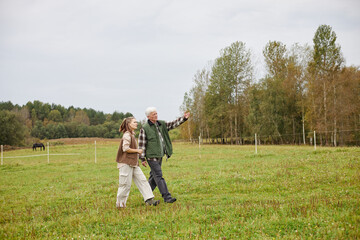 Senior Caucasian man and young adult Caucasian woman walking together in grassy field, man pointing...