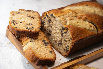 Freshly baked banana bread is sliced and displayed on a wooden cutting board. The bread has a golden-brown crust and chocolate chips, perfect for a cozy snack at home.