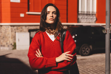 Confident young woman wear red sweater posing with crossed arms against red building standing in warm sunlight. Natural light fashion portrait with stylish attitude and relaxed posture.