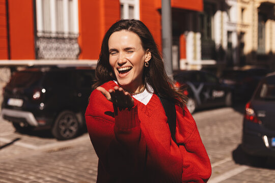 Portrait close up brunette long hair woman send blow air kiss, wear red sweater. Woman outdoors blowing kiss on city street evokes warm expression featuring a casual outfit. 