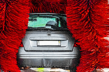 Rear view of silver car in automatic self service wash with red rotating brushes, water spray and...
