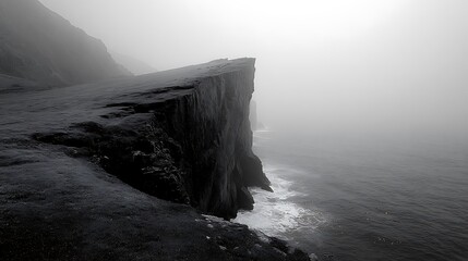 Dramatic black and white image of a steep coastal cliff face meeting the ocean in foggy conditions