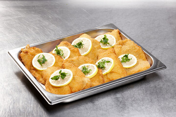 A tray is filled with golden fried fish fillets, each topped with lemon slices and fresh cilantro, placed in a busy kitchen setting, evoking a vibrant culinary atmosphere