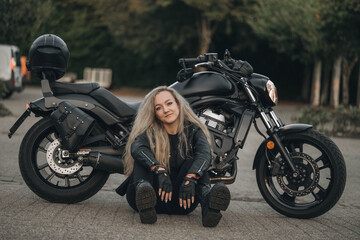 Obraz premium Smiling woman in black leather jacket sitting on the ground near a motorcycle, enjoying freedom and adventure outdoors.