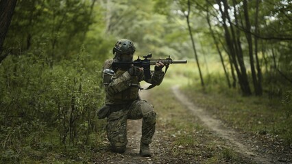 Camouflaged army soldier with AR15 carbine on a mission. Kneeling and taking aim with the rifle. Military recon