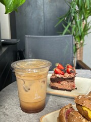Iced coffee, chocolate strawberry cake, and sandwich buns with fillings on a gray table in a cafe. Background includes plants and a chair.