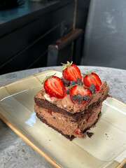 A slice of chocolate cake with strawberries and chocolate chunks on a beige plate, placed on a grey marble table in a cafe setting.