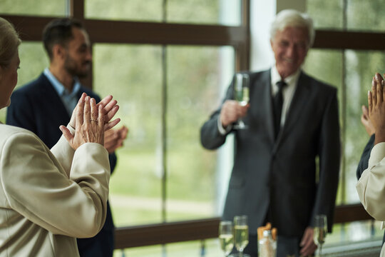 Senior Caucasian man holding champagne glass standing near window, smiling while diverse group of adults clapping and celebrating during formal gathering or special event