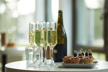 Closeup showing four champagne flutes filled with sparkling wine standing on round table near bottle and dessert plate, blurred background suggesting indoor celebration or gathering