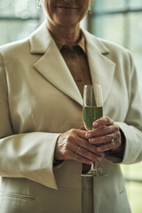 Middle aged Caucasian woman holding champagne flute with green beverage, standing indoors with hands visible, wearing formal suit, smiling slightly, not looking at camera