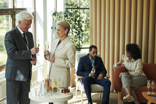 Senior Caucasian man and senior Caucasian woman standing and holding champagne glasses, talking near table with drinks, young Black woman and young Middle Eastern man sitting and drinking - Powered by Adobe