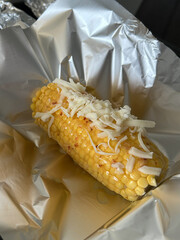 Corn on the cob with melted butter, chili flakes, and grated cheese placed on aluminum foil, ready for baking or grilling.