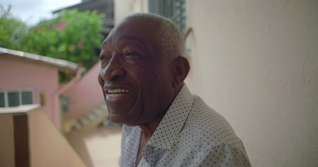 One Elderly Black man sitting outdoors on porch, laughing warmly, exuding joy and positivity, tropical residential surroundings with pink walls and greenery in background