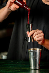 Bartender pouring red liquor into jigger above cocktail shaker on green bar counter. Close-up of drink preparation in professional bar setting.