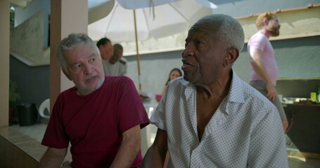 Obraz premium Two elderly men sitting and chatting over drinks in a casual outdoor setting, relaxed and enjoying friendly conversation with a barbecue event in the background