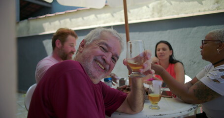 Elderly man smiling and taking a sip of beer from a glass at a backyard barbecue, embodying joy and connection during a lively gathering with family and friends