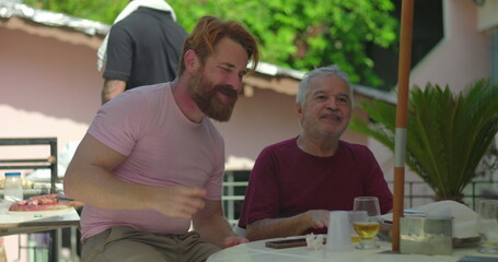 Elderly man joyfully engaging with a younger man in an outdoor barbecue setting, smiling and dancing together in a moment of celebration and shared happiness