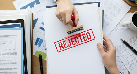 Hands stamping "REJECTED" on a document amidst paperwork and a coffee cup on a desk.