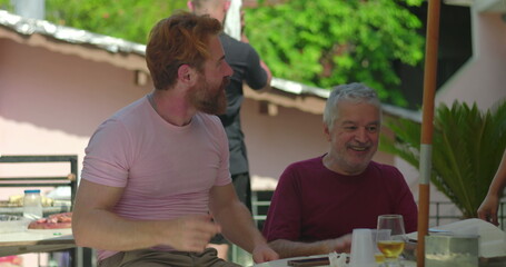 Elderly man joyfully engaging with a younger man in an outdoor barbecue setting, smiling and...