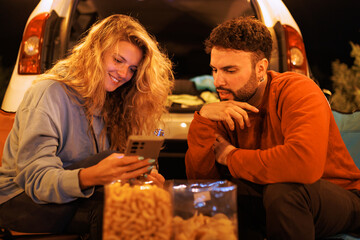 Friends enjoying snacks and sharing moments while sitting by a car at night, captured during a cozy outdoor gathering in a serene location