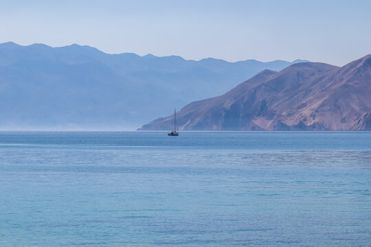 A solitary sailboat glides across the calm, blue Adriatic Sea on a hazy day near Baska, Croatia, with layers of distant, majestic mountains creating a serene and peaceful coastal landscape. - Powered by Adobe