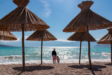 A mother and child are silhouetted against the sparkling Adriatic Sea on a pebble beach with straw umbrellas, looking towards the mountains from the coast of Baška, Krk island, Croatia.