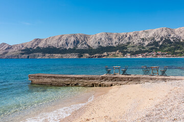 Two empty tables with chairs sit on a stone pier by a tranquil sandy beach, offering a stunning view of the coastal town and rocky mountains of Baška on Krk island, Croatia.