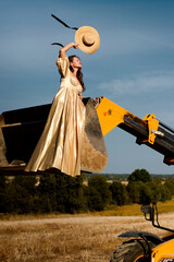 Elegant woman in a gold dress stands confidently on a yellow excavator with a straw hat, overlooking harvested fields under a blue sky, blending elegance with rustic charm. © Oleksandr