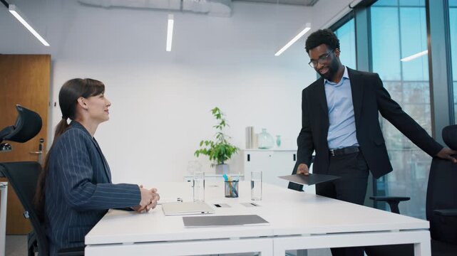 Smiling African American man in suit entering office, greeting Caucasian female HR manager during interview. Confident job candidate holding folder with resume in professional, modern meeting room.