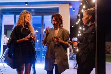 Three women having a conversation while enjoying drinks at an outdoor evening gathering.