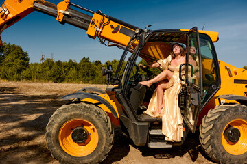 Elegant woman in a flowing golden gown playfully relaxes inside a yellow telescopic handler against the backdrop of a sunny construction site, creating an intriguing blend of fashion and industry.