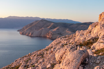 Sunlit rocky terrain stretches down to calm blue waters, with steep cliffs and the island of Prvić illuminated by early morning light, seen from high above Baška, Krk, Croatia.