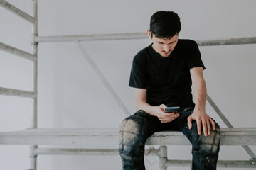 A young worker sits on scaffolding with a smartphone in his hands.