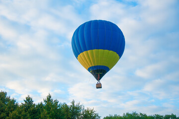 Fototapeta premium Blue and yellow hot air balloon floating above green trees under a partly cloudy sky. A serene aerial scene blending adventure and national colors