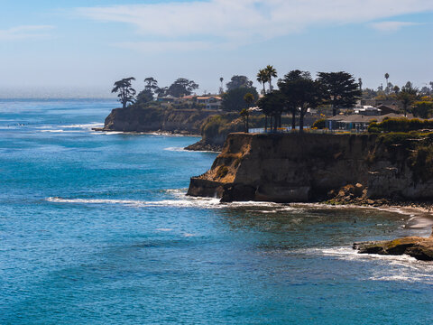 Aerial view of Capitola, California, showing cliffs with houses and trees, turquoise Pacific waters, small waves, and a clear sky with scattered clouds.