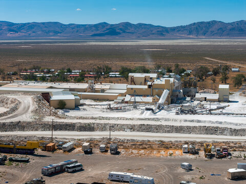 Aerial view of a salt mining site in Nevada with beige buildings, machinery, and white salt piles, set against a desert and mountain backdrop.