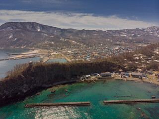 Naklejka premium Aerial view of a coastal town with turquoise waters, harbor structures, and snow-dusted mountains in the background