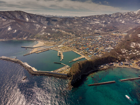 Aerial view of a coastal fishing town with scenic harbor, turquoise waters, and snow-capped mountains in the background