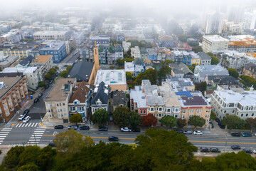 Obraz premium Aerial view of colorful Victorian style houses in San Francisco with a tree lined street, parked cars, and a church steeple in the foggy background.