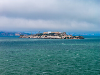 Fototapeta premium Alcatraz Island in San Francisco Bay features historic prison buildings, rugged terrain, and a watchtower, surrounded by turquoise waters under a cloudy sky.