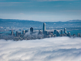 Aerial view of San Francisco featuring the skyline with Salesforce Tower, other skyscrapers, fog over the city, and the San Francisco Bay in the background.