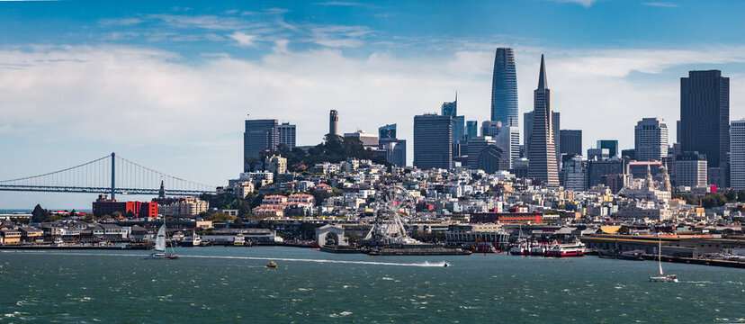 Aerial view of San Francisco featuring the Transamerica Pyramid, Salesforce Tower, waterfront activity, ferris wheel, and partial Golden Gate Bridge.
