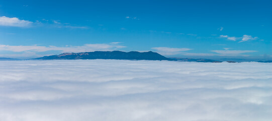 A mountain range rises above a thick cloud layer under a clear blue sky, showcasing the natural...