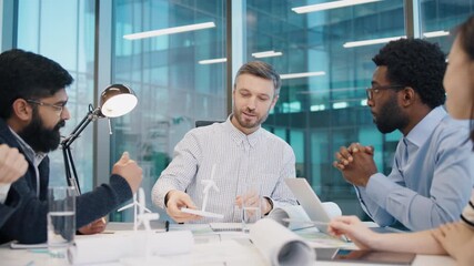 Caucasian male engineer explaining wind turbine model during team discussion with diverse colleagues at conference table. Professionals brainstorming renewable energy project in bright office. - Powered by Adobe