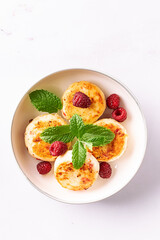 Cottage cheese pancakes with honey, raspberry and mint leaves on white ceramic plate on white marble table top view. Healthy homemade dessert.