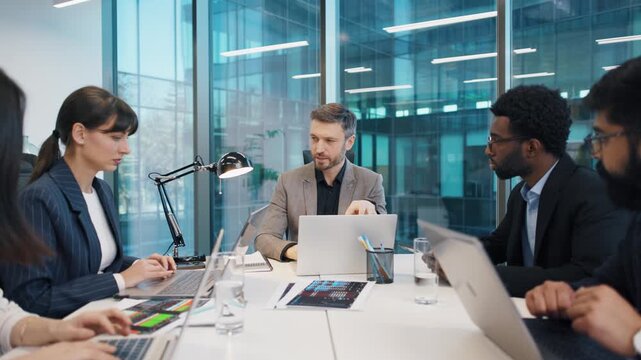 Business team working at table with laptops and charts, leader pointing while discussing strategy. Focused professionals in formal attire engaged in project planning in modern office.