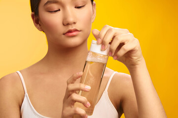 asian woman holds a clear bottle of liquid, inspecting its contents with a calm expression in a bright studio. vibrant yellow background enhances a fresh, contemporary lifestyle mood and minimalist